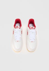 Fehér Nike Air Force 1 sportcipők piros kiegészítőkkel, sima bőr felsőrésszel, perforált orral és fehér fűzővel. Gumi talp.