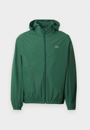 Green lightweight jacket with a hood, zip closure, elastic cuffs, and an elastic waistband. Features a small logo on the chest.
