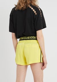 T-shirt nera con aperture sulle spalle e accessori decorativi, abbinata a pantaloni corti gialli vivaci con vita elastica nera e strisce laterali.