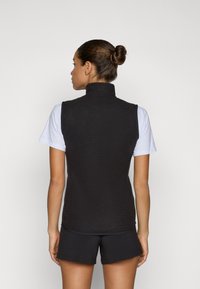 Zwarte mouwloze vest met een hoge kraag, gestructureerde stof, getailleerd ontwerp en zijzakken, gedragen over een wit t-shirt en zwarte shorts.