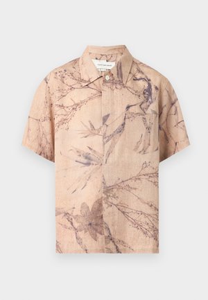 Personne debout de profil portant une chemise beige ample à manches courtes à motifs floraux, un jean large bleu clair et des chaussures noires sur un fond uni.