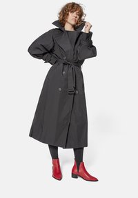 Cappotto lungo nero con colletto oversize, vita con cintura e dettagli in bottone, abbinato a stivaletti rossi fino alla caviglia e leggings neri. Texture liscia.