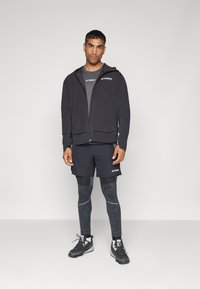Chaqueta negra con cremallera y capucha, camiseta gris, pantalones cortos negros, leggings estampados y zapatillas negras con detalles reflectantes. Marca Terrex visible.