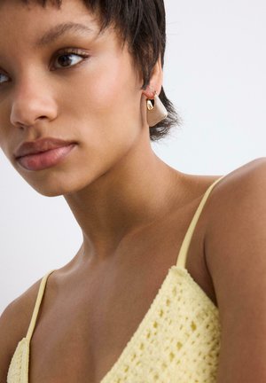Mlada žena s kratkom tamnom kosom nosi žuti heklanog top i velike bež i zlatne naušnice, gleda lagano ulijevo.