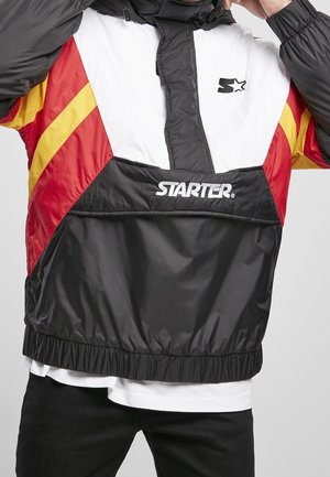 Personne portant une veste coupe-vent Starter noire, blanche, rouge et jaune avec un logo sur la poitrine et la poche avant, ajustant la capuche.