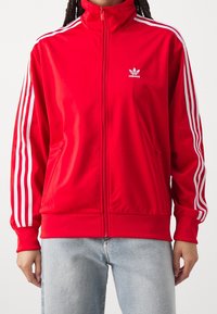 Czerwona kurtka zapinana na zamek wykonana z gładkiego materiału, z białymi trzema paskami na rękawach i haftowanym logo na piersi.