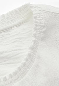 Witte katoenen top met een textuurpatroon, featuring een ruches halslijn. De stof lijkt zacht en lichtgewicht, wat het delicate ontwerp versterkt.