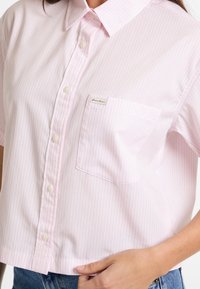 Camicetta con bottoni tagliata in rosa chiaro con righe bianche verticali. Presenta una tasca sul petto e un'etichetta con il marchio sulla tasca. Tessuto morbido.