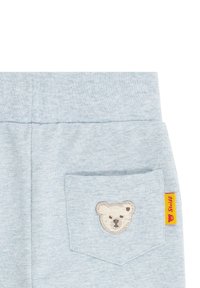 Hellblaue Kinderhose aus Baumwolle, mit einem gestickten Bärenkopf auf der Rückentasche und einem beigefügten gelben Markenetikett.