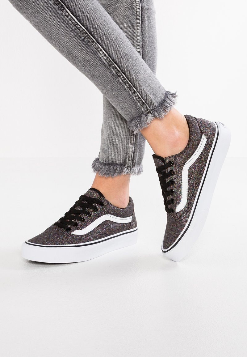 Pieds chaussés de baskets noires pailletées avec des bandes blanches sur les côtés et des lacets noirs, associées à un jean gris à ourlet effiloché.