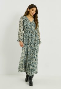 Robe longue à motifs floraux en vert et crème, avec un col en V, des manches longues et une taille à cordon de serrage. Comprend une jupe à volants et des poignets à volants.