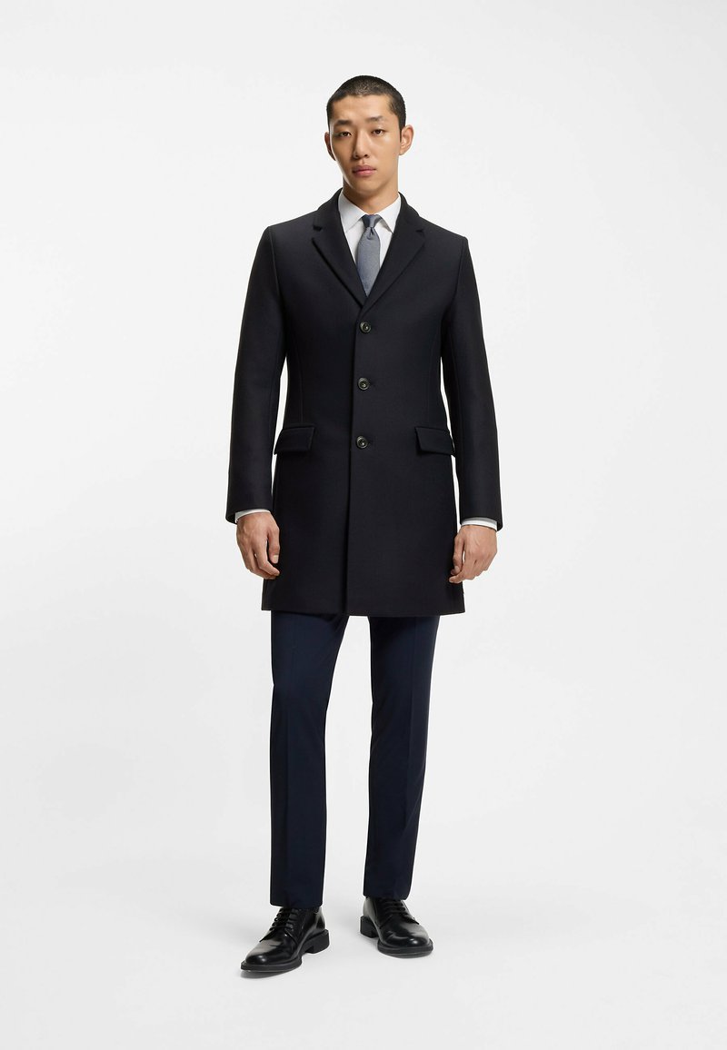 Cappotto navy su misura con cinque bottoni, due tasche frontali e revers strutturati, abbinato a pantaloni navy coordinati e scarpe nere stringate.