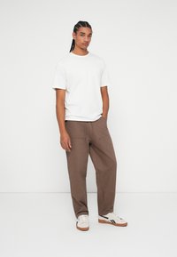 T-shirt bianco in cotone abbinato a pantaloni marroni larghi con dettagli di coulisse e tasche. Modello con sneakers chiare.