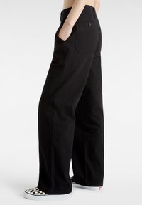 Zwarte wijde broek met hoge taille, voorzien van zijzakken en een achterzak met knoop; gedragen met geruite instappers.