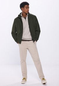 Olive green zip-up jacket over a light beige sweater, paired with light beige pants and white sneakers with yellow laces and accents.