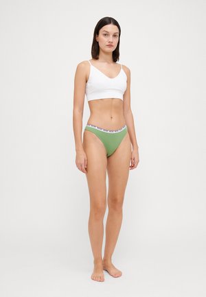 Vit bralette med tunna band, parat med gröna bikiniunderbyxor med ett mönstrat midjeband. Modellen står barfota mot en ljus bakgrund.