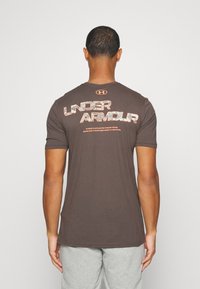 Brun bomulls-T-shirt med en stor "UNDER ARMOUR" logotyp i en kontrasterande mönster på baksidan, rund hals och korta ärmar.