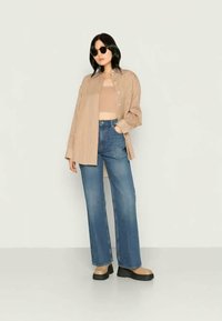 Camisa oversized bege riscada, top de alças castanho claro, jeans azuis e sapatos de plataforma pretos e chunky. Modelo usa óculos de sol redondos.