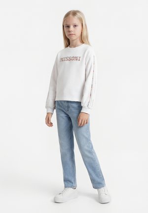 Jeune fille debout, portant un sweat-shirt blanc avec un logo à motif et des rayures sur les manches, un jean bleu clair et des baskets blanches.