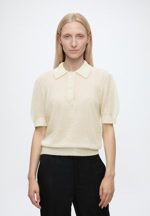 Femme aux cheveux blonds raides porte une chemise beige clair à manches courtes avec col et un pantalon noir, debout devant un fond blanc uni.