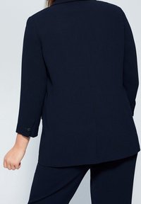 Námořnicky modré sako na míru s tříčtvrtečními rukávy a knoflíkovými manžetami. Hladká látka se rovnými liniemi a minimálním zdobením na zadní části.