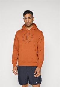 Rustfarvet hættetrøje lavet af blødt stof, med en kængurulomme, snøre i hætten og et cirkulært logodesign på forsiden.