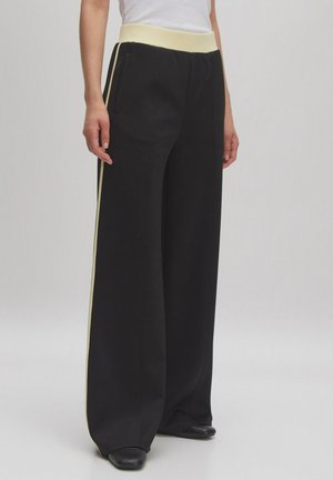 Schwarze weite Hosen verfügen über einen cremefarbenen, elastischen Bund und cremefarbene Seitestreifen. Sie bestehen aus einem glatten, maßgeschneiderten Stoff und haben seitliche Taschen.