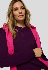 Lila gestricktes Poloshirt mit geknöpftem Kragen, kombiniert mit einem rosa Hoodie, der über die Schultern gelegt ist. Glatte Textur, eng anliegendes Design.