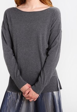 Femme portant un pull gris charbon à manches longues avec un col bateau et une jupe plissée métallique, mains jointes devant.