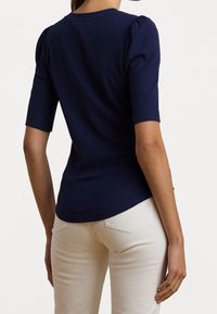 Žena v tmavě modrém ribbeném topu s krátkým rukávem a světle béžových kalhotách, zobrazená zezadu na jednobarevném pozadí.