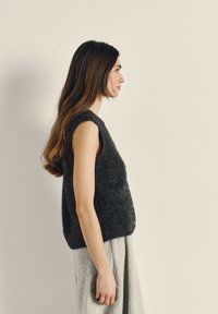 Grijs mouwloos gebreid vest met een textuurafwerking en open zijkanten. Gepaard met een lichtgrijze rok, gedragen door een persoon van opzij.