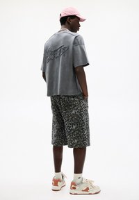 Grå bomulds t-shirt med broderet logo på ryggen, parret med knælange shorts med leopardprint. Modellen har en lyserød kasket og multikolore sneakers på.