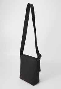 Černá crossbody taška vyrobená z odolné látky, s obdélníkovým tvarem, nastavitelným popruhem a zipovým uzávěrem na vrchu.