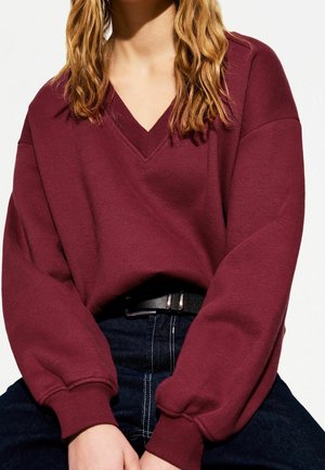 Femme portant un pull col en V bordeaux avec des manches bouffantes et un jean foncé, les mains posées sur les genoux, cadrée au niveau du cou et de la taille.