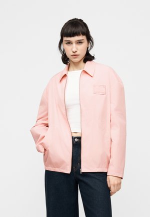 Νεαρή γυναίκα που φοράει ένα τζάκετ ανοιχτό ροζ με φερμουάρ πάνω από λευκό cropped τοπ και σκούρο τζιν, στέκεται με το ένα χέρι στην τσέπη.