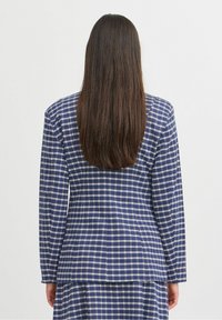 Personne aux longs cheveux bruns portant un blazer à carreaux bleu et blanc avec une jupe assortie, vue de dos sur un fond uni.