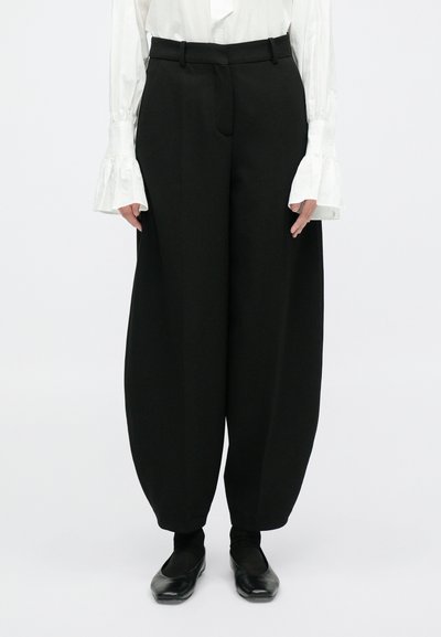 Lässige schwarze Hosen haben eine glatte Textur, einen hohen Bund und Seitentaschen, kombiniert mit schwarzen flachen Schuhen und weißen, voluminösen Ärmeln.