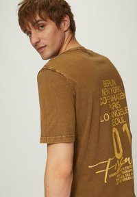 Olijfgroene t-shirt met korte mouwen en een comfortabele pasvorm, met op de achterkant gedurfde gele opdruk en een ronde halslijn.