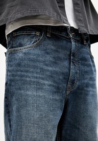 Primer plano de unos vaqueros de mezclilla azules gastados con costuras naranjas y una camisa oscura parcialmente metida por dentro de una camiseta blanca.