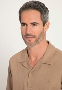 Chemise beige à manches courtes avec col cranté, comportant un logo brodé discret sur la poitrine et un tissu léger et texturé.