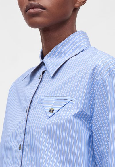 Gros plan d'une personne portant une chemise boutonnée à rayures verticales bleues et blanches, avec un col pointu et une poche poitrine triangulaire boutonnée.