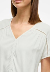 Blusa beige chiaro con scollo a V, maniche corte e cuciture decorative lungo le cuciture. Tessuto morbido e fluido con una texture liscia.