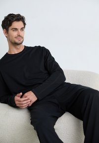 Chemise noire à manches longues et pantalon assorti sur une figure assise. Tissu doux avec des détails de couture minimaux, sur fond clair.