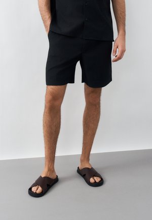 Άνδρας φοράει μαύρα κοντά παντελόνια με υφή και ταίριαστο πουκάμισο, στέκεται ξυπόλητος σε μαύρα και καφέ σανδάλια slide σε γκρι πάτωμα.