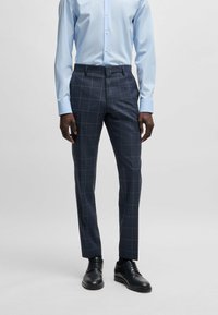 Pantaloni a quadri navy con una vestibilità su misura, realizzati in un tessuto liscio. Indossati con una camicia button-up azzurro chiaro e scarpe eleganti nere.