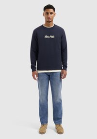 Marineblauw sweatshirt met wit logo, gedragen over een crèmekleurig shirt. Gecombineerd met lichtblauwe denim jeans en tan schoenen. Casual pasvorm en ontwerp.