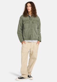 Chaqueta de pana verde con dos bolsillos en el pecho, llevada sobre una camisa gris; pantalones beige de corte holgado y zapatillas negras completan el atuendo.