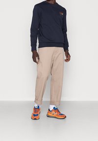 Námořnicky modrá mikina, khaki kalhoty a vícebarevné tenisky (oranžové, modré, černé) nošené s bílými ponožkami; ležérní střih s viditelnými švy.