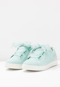 Sneaker in suede verde menta con lacci in nastro trasparente, dettaglio metallico sul tallone e suola in gomma bianca. Texture liscia e design con punta rotonda.