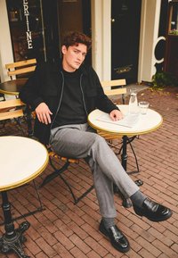 Black jacket over a black shirt, gray trousers, and black shoes. Sitting at a round table on a brick patio with glassware and a newspaper.